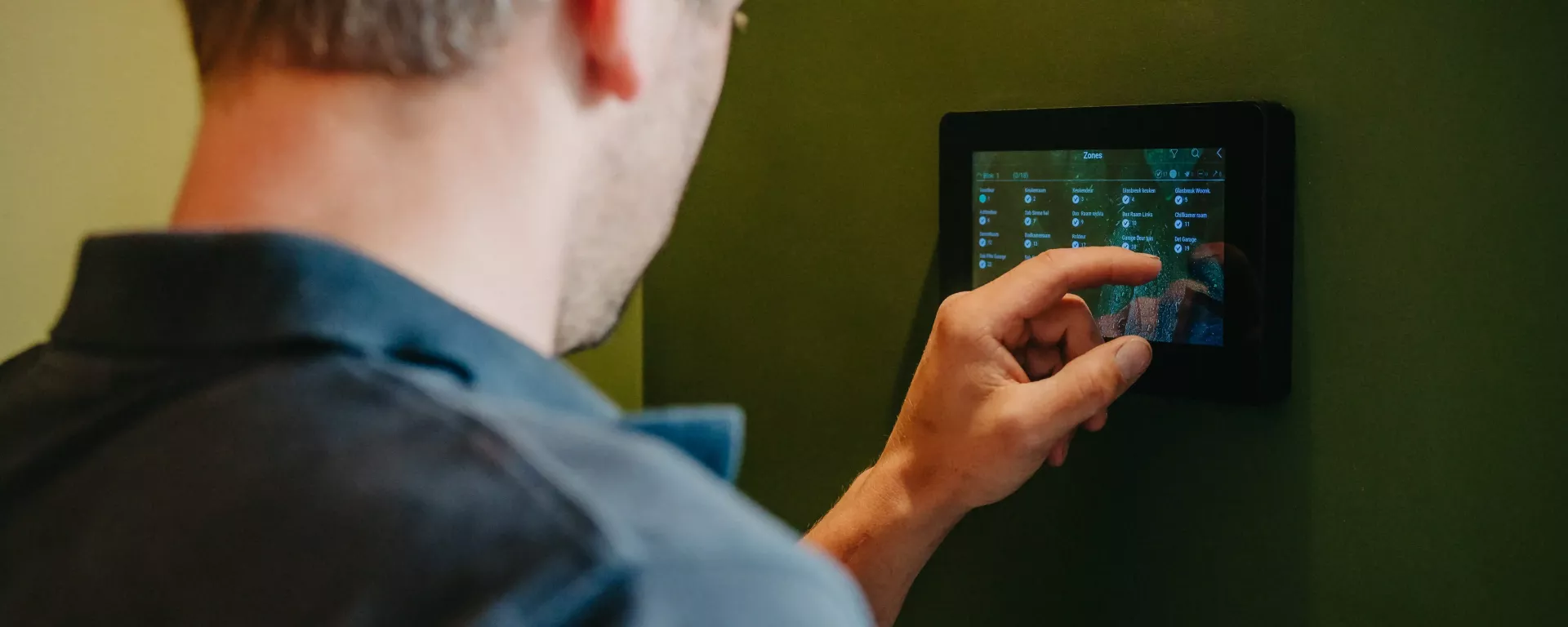 Technicus bedient een muur gemonteerd touchscreen voor het instellen van een domotica- of klimaatbeheersingssysteem, tegen een groene achtergrond.