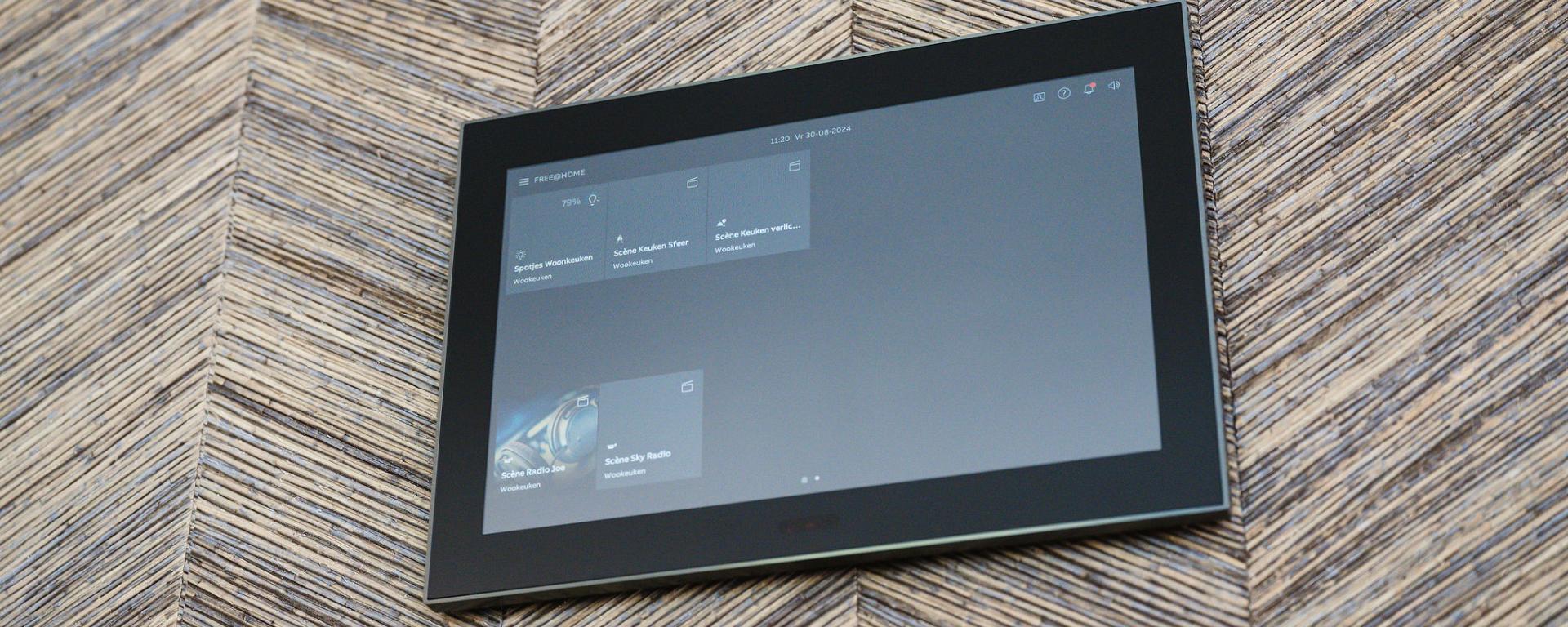 Wandgemonteerd touchscreen voor domotica-bediening met een strak ontwerp, geïnstalleerd tegen een decoratieve wand met een visgraatstructuur.