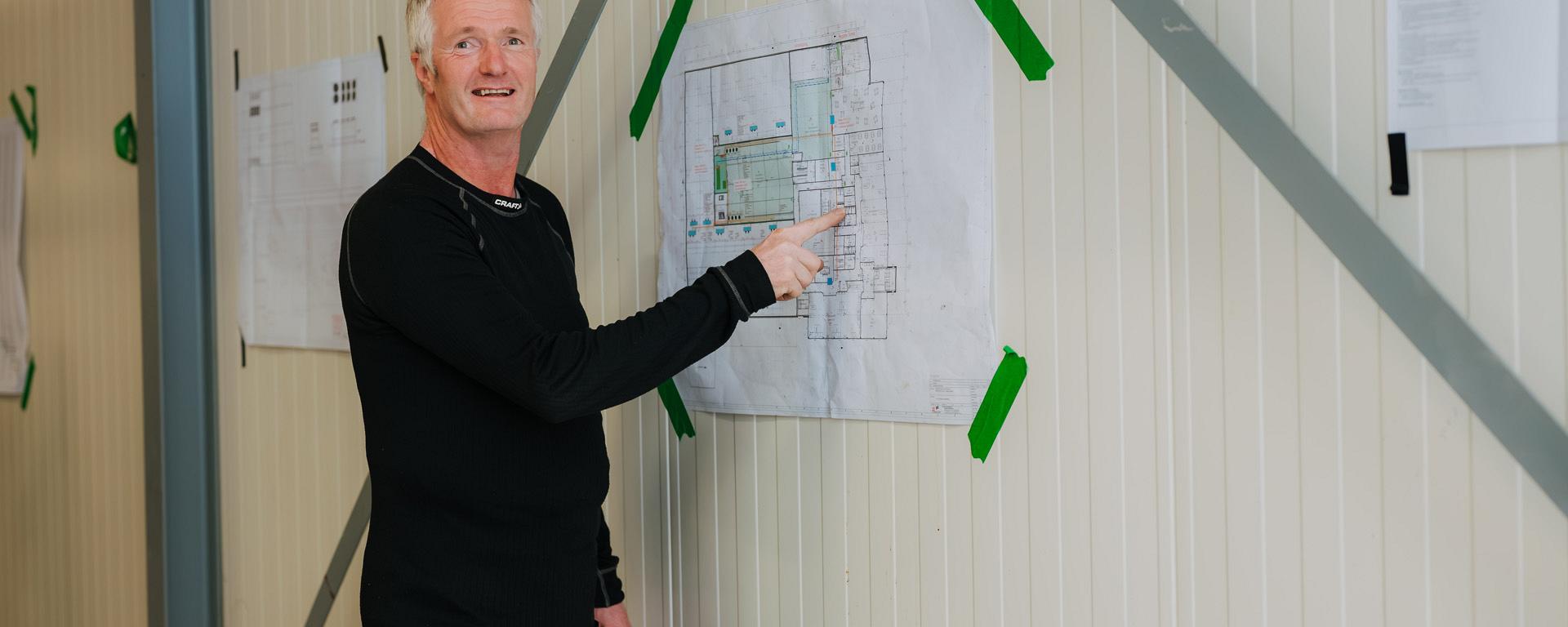 Een man wijst naar een bouwtekening op een wand in een binnenruimte.