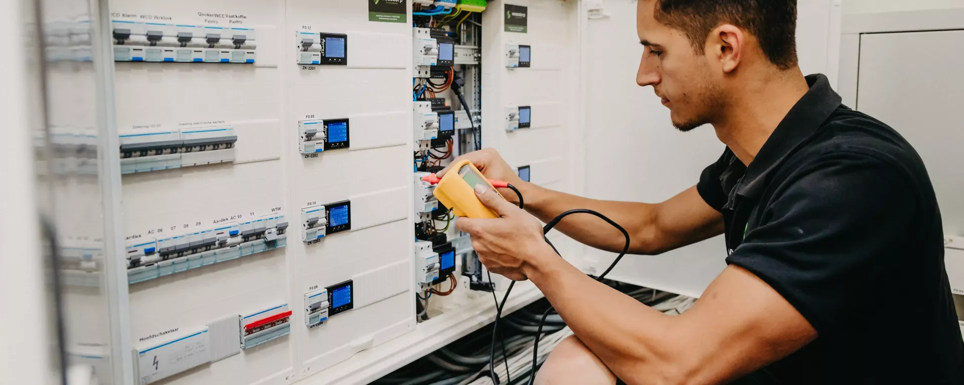 Technicus voert een meting uit in een geavanceerde elektrische verdeelkast, met meetapparatuur in de hand en zicht op bedrading en schakelaars in een technische ruimte.