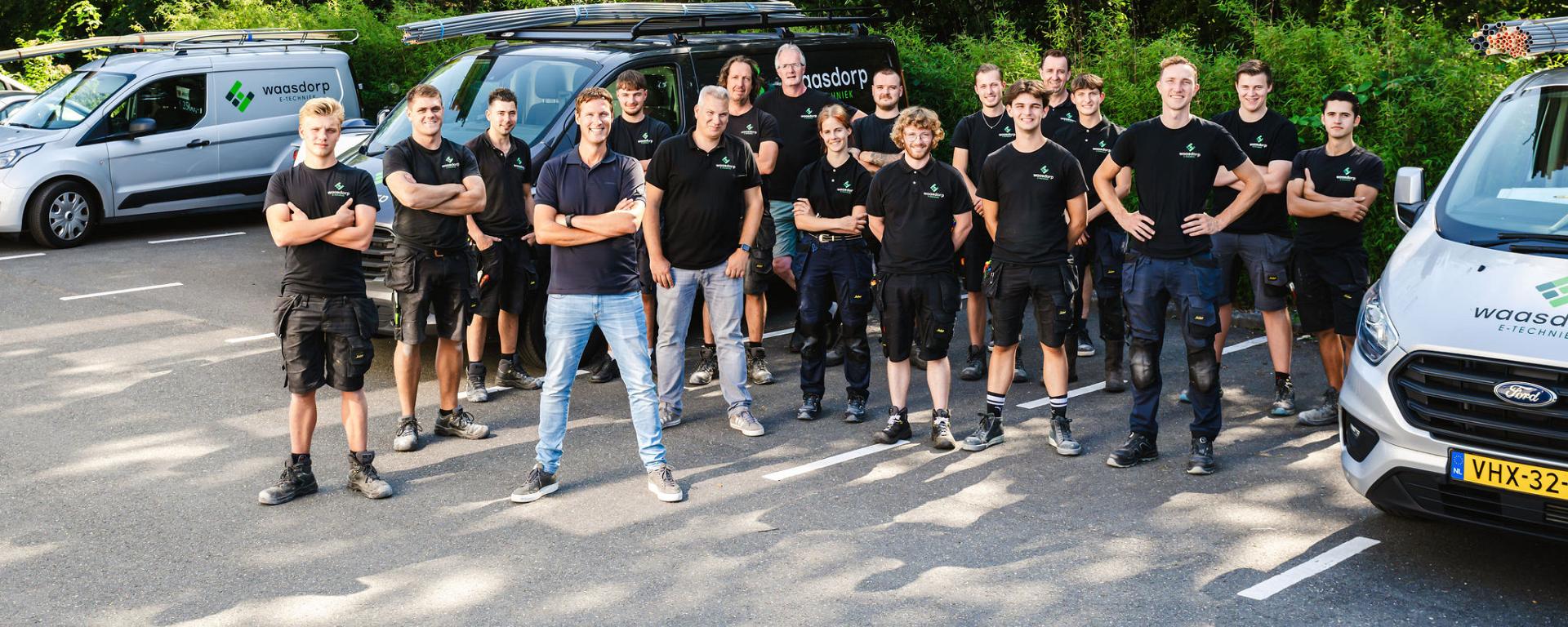 Een team van monteurs poseert in werkkleding voor bedrijfswagens met ladders, in een groene parkeeromgeving, klaar voor hun werkzaamheden.