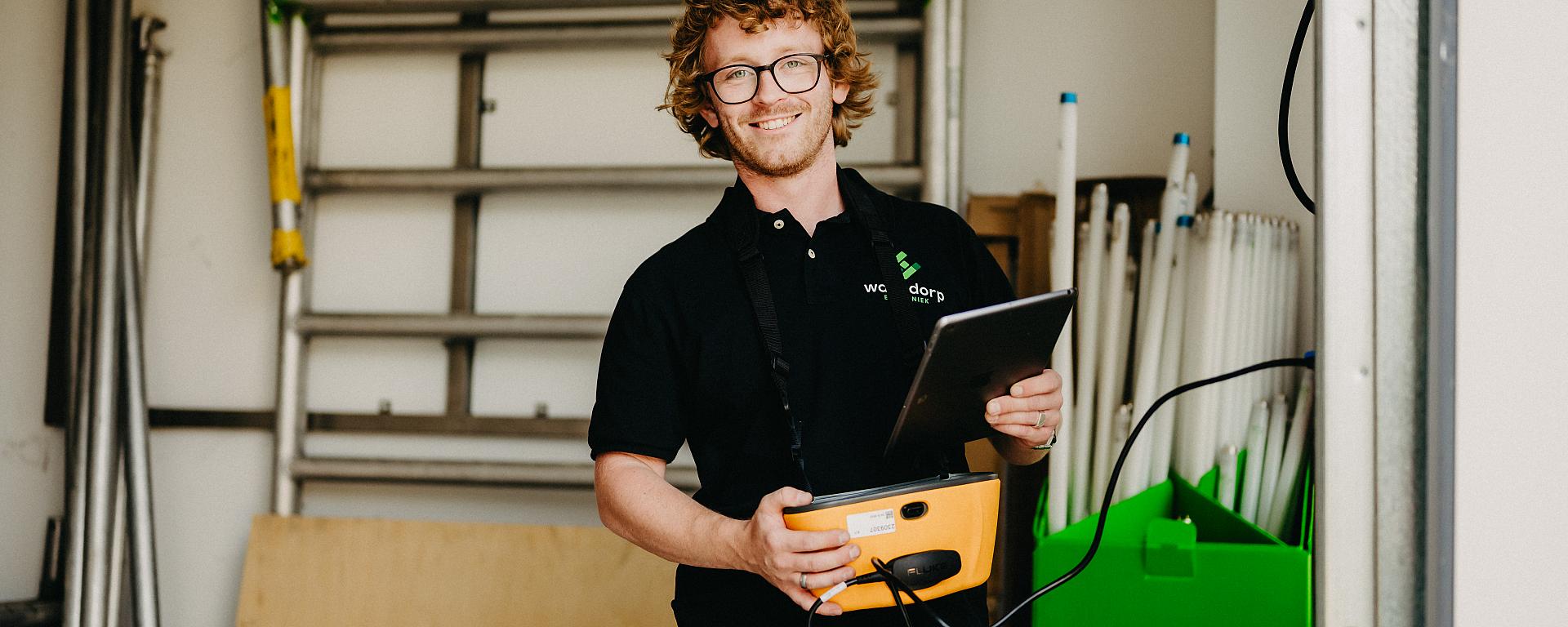 Monteur van Waasdorp met meetapparatuur en tablet in werkruimte, klaar voor elektrotechnische controle.