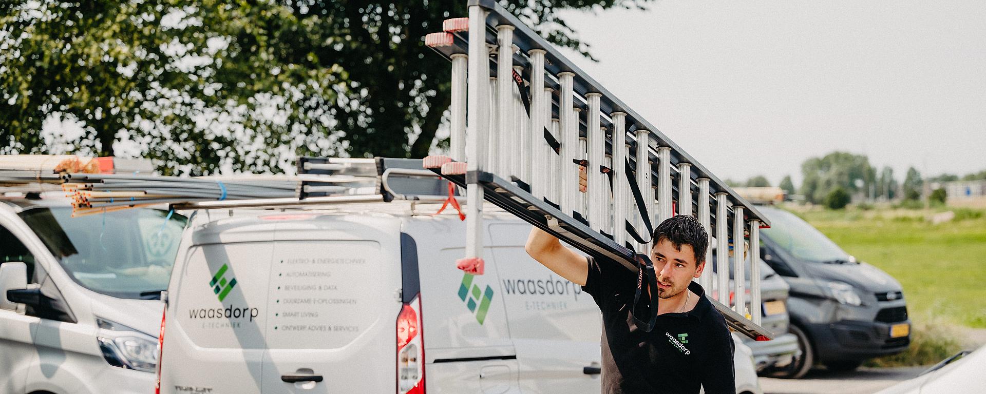 Monteur van Waasdorp tilt ladder naast bedrijfsbus op parkeerterrein met servicewagens.