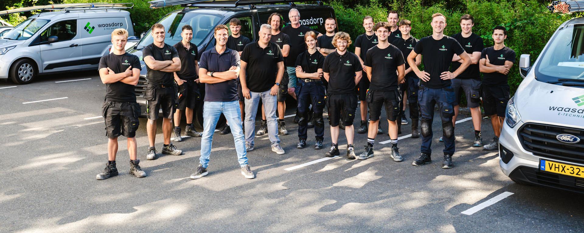 Een team van monteurs poseert in werkkleding voor bedrijfswagens met ladders, in een groene parkeeromgeving, klaar voor hun werkzaamheden.