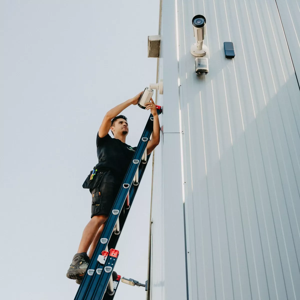 Monteur op een ladder installeert een beveiligingscamera aan de buitenkant van een gebouw met een metalen gevel, onder een heldere blauwe hemel.