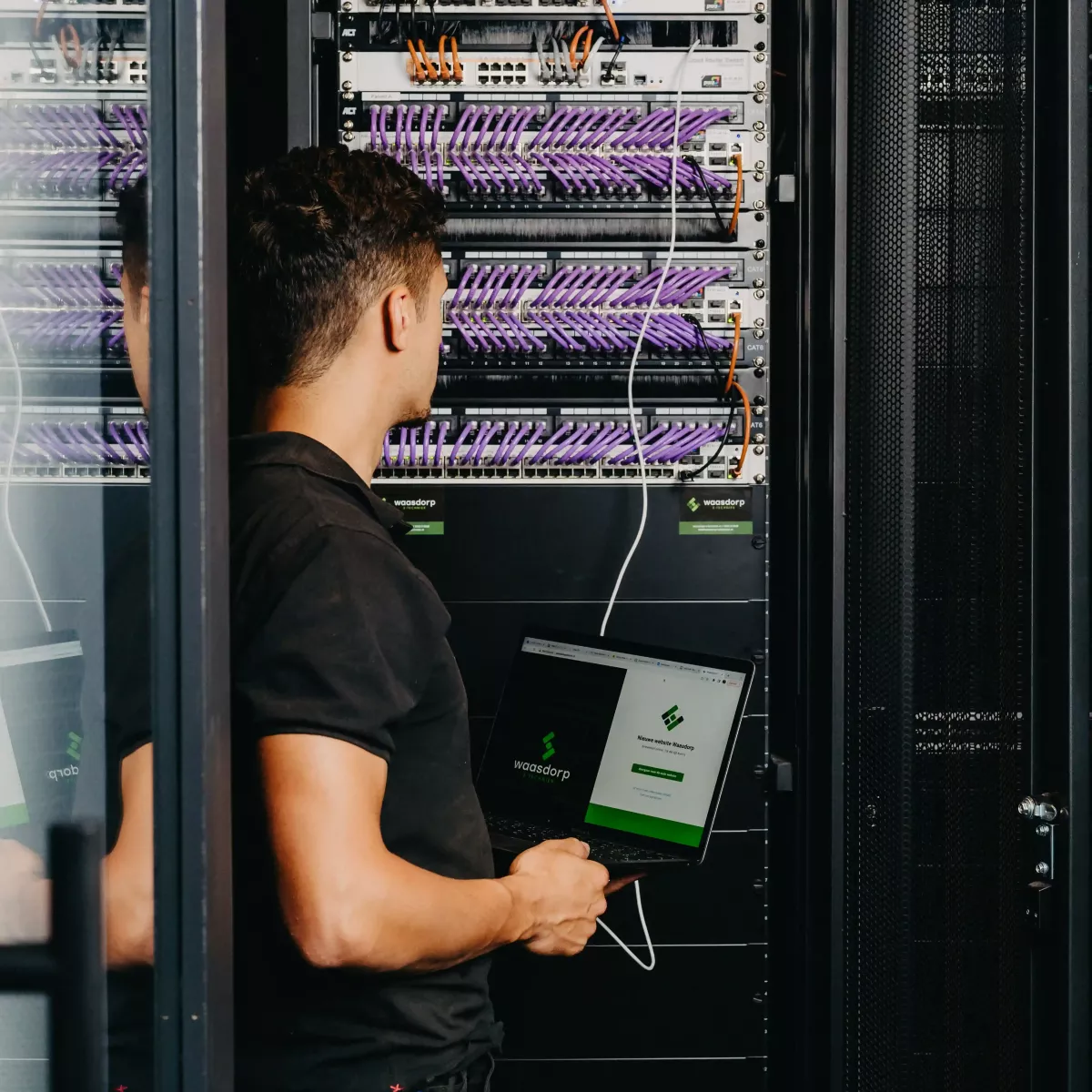Technicus werkt in een serverruimte met een laptop in de hand, omringd door serverkasten gevuld met netjes georganiseerde paarse netwerkbekabeling.