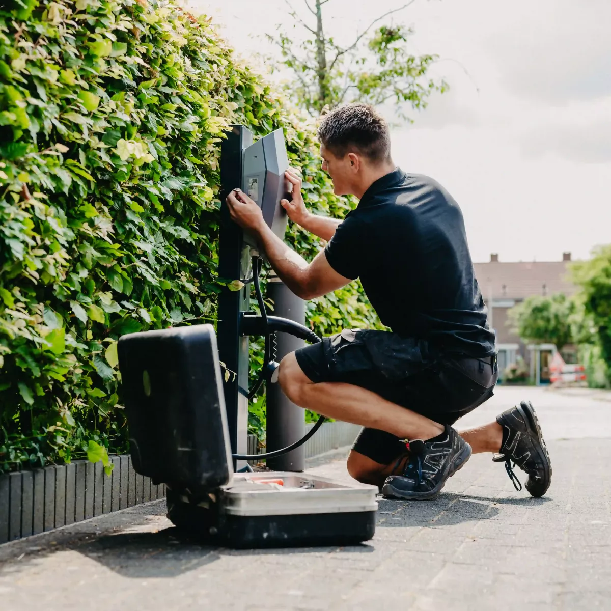 Monteur installeert een laadpaal voor elektrische voertuigen langs een groene haag, met een open gereedschapskoffer op de grond en een woonwijk op de achtergrond.