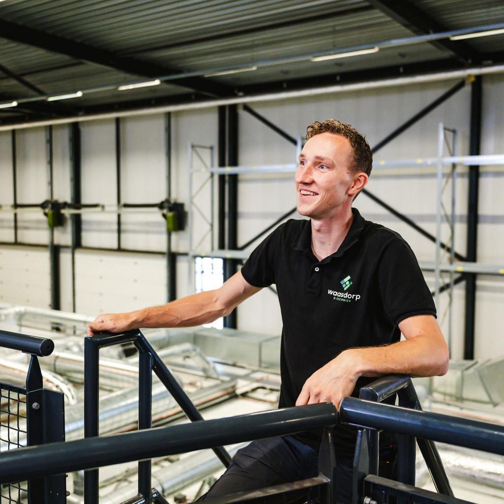 Lachende technicus in Waasdorp-bedrijfskleding leunt op een reling in een moderne industriële hal, met ventilatiesystemen en hoge plafonds op de achtergrond.