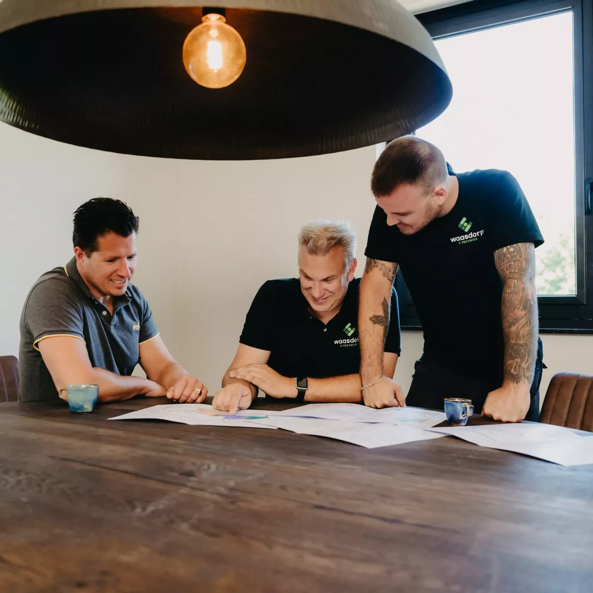Drie mannen bespreken bouwtekeningen aan een houten tafel in een moderne vergaderruimte, verlicht door een grote hanglamp, met koffie op tafel.