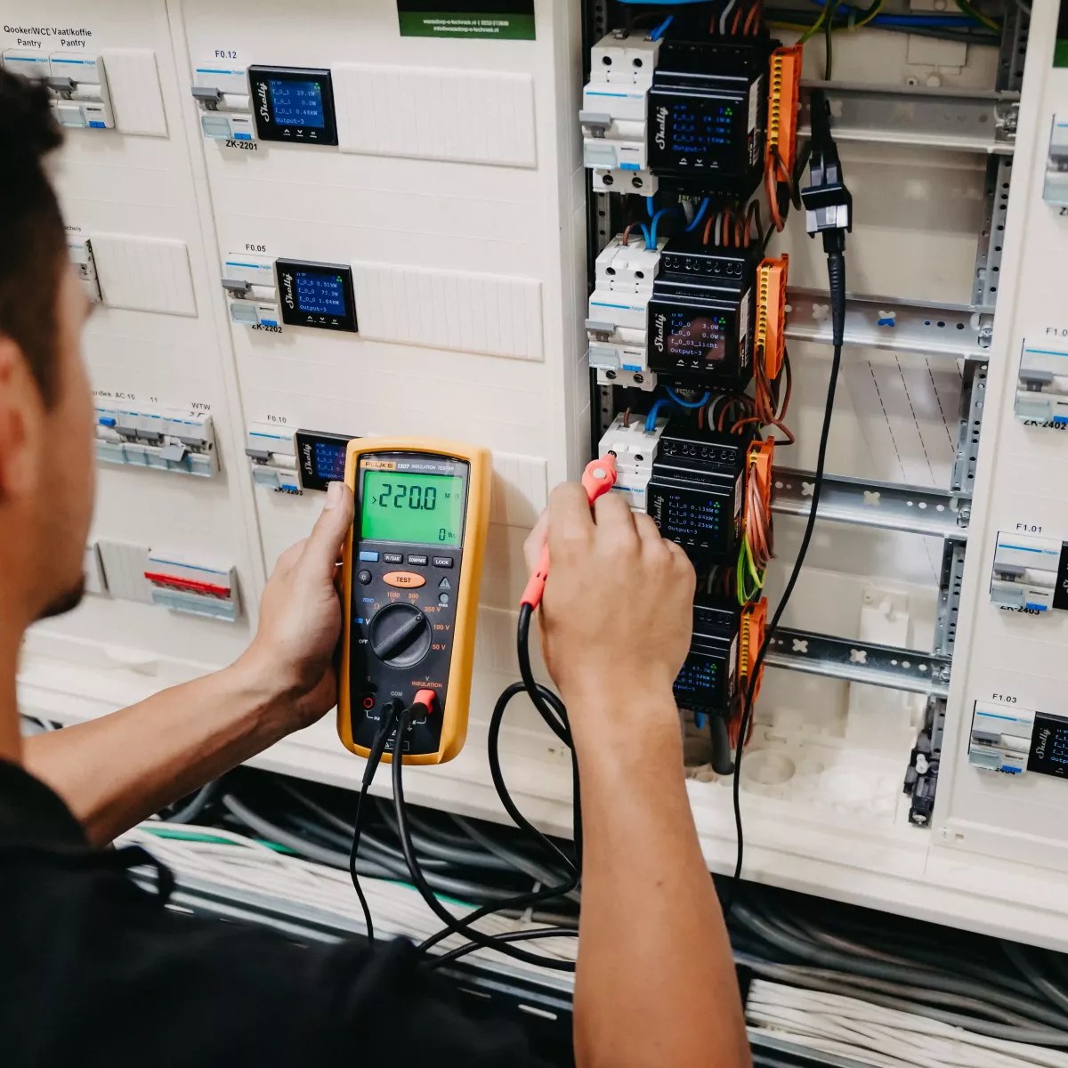 Technicus voert spanningsmeting uit met een multimeter op een elektrische installatiekast, waarbij een display 220 volt aangeeft en bedrading zichtbaar is.