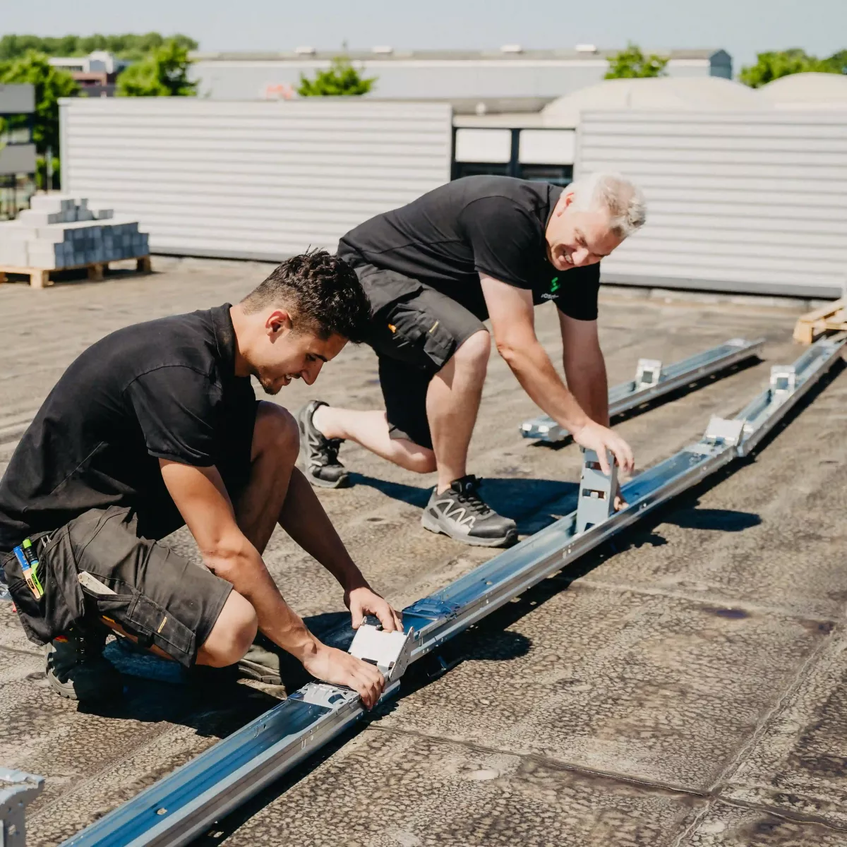 Twee monteurs bevestigen montagerails op een plat dak als voorbereiding voor de installatie van zonnepanelen, met een achtergrond van bedrijfspanden en groen.