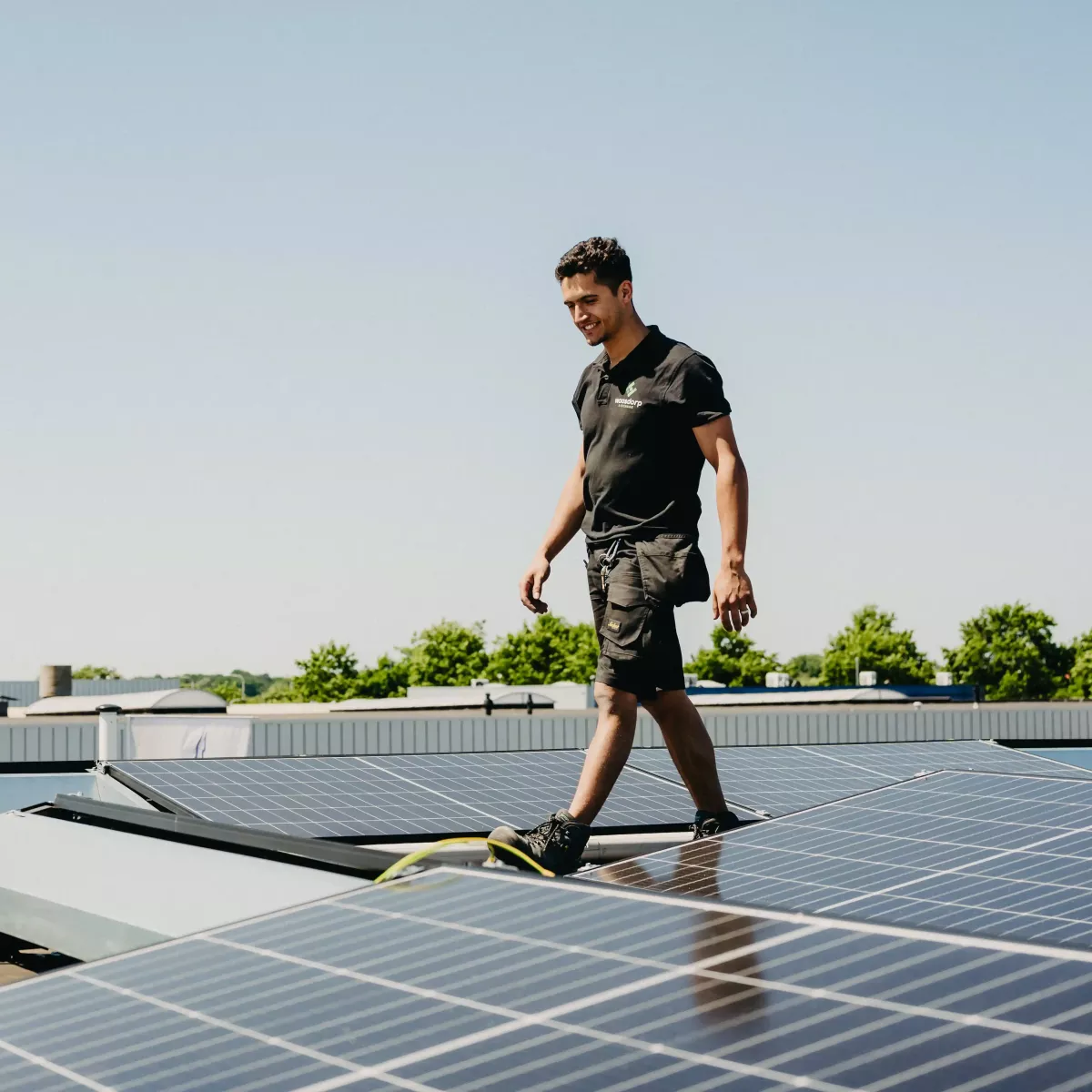 Technicus loopt over een plat dak met geïnstalleerde zonnepanelen, onder een heldere blauwe lucht, omgeven door groene bomen en industriële gebouwen.