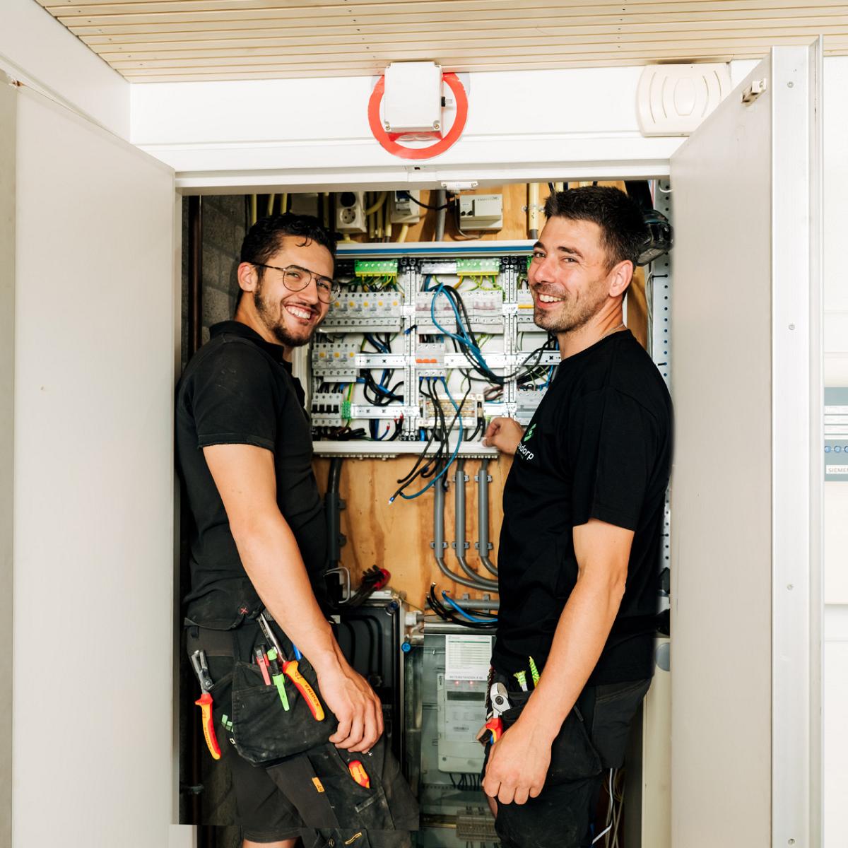 Twee elektriciens staan lachend voor een geopende schakelkast met elektrische bedrading en installatiemateriaal.