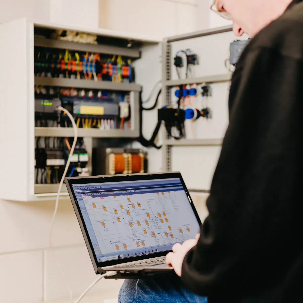 Technicus werkt met een laptop aan een geopende schakelkast, met een zichtbaar digitaal schema op het laptopscherm en bedrading in de kast.
