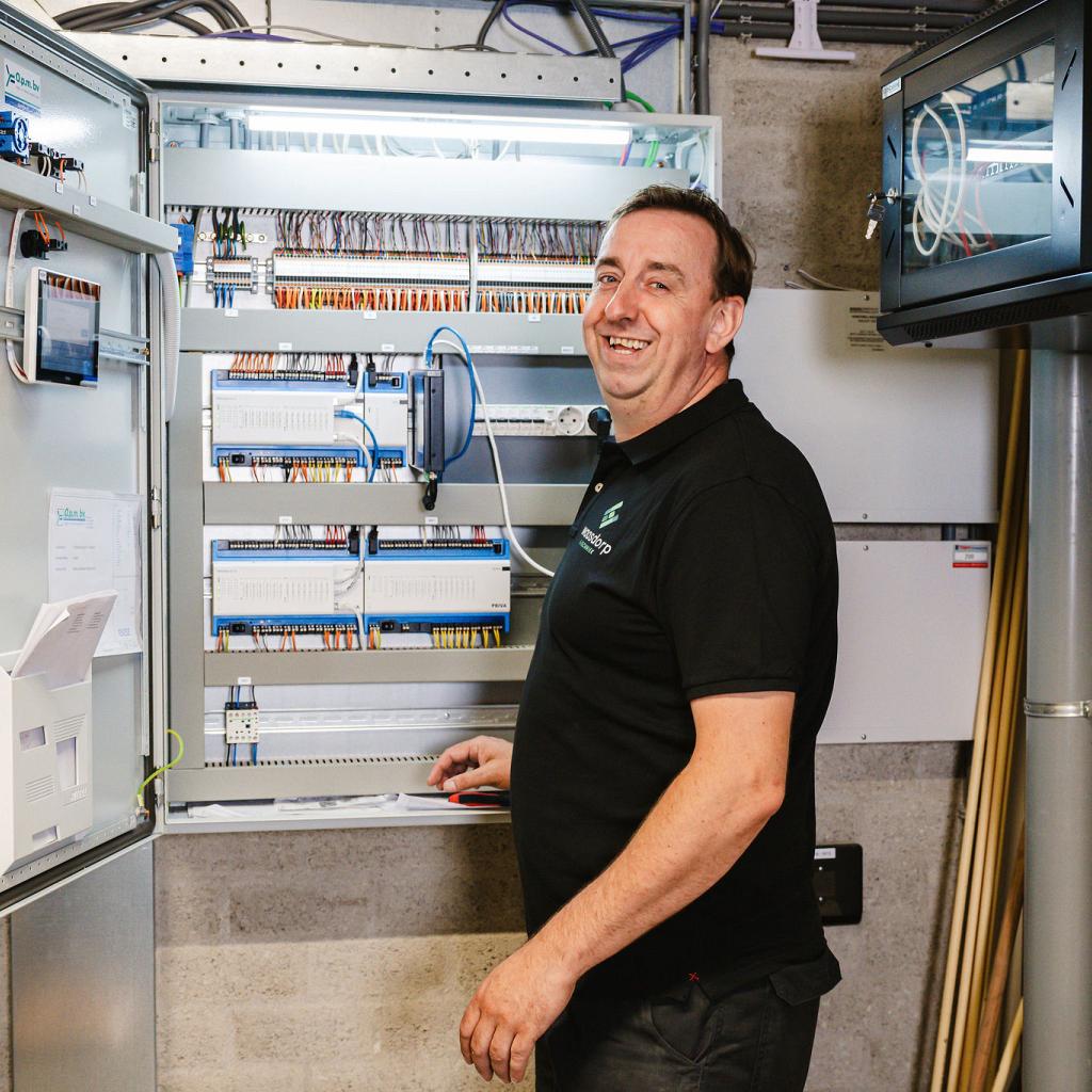 Lachende technicus werkt aan een geopende schakelkast met overzichtelijke bedrading en elektronische componenten in een technische ruimte.