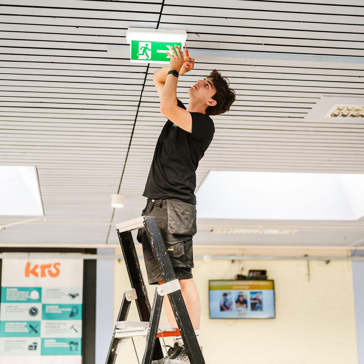 Monteur plaatst een nooduitgangsbord aan het plafond terwijl hij op een trap staat.