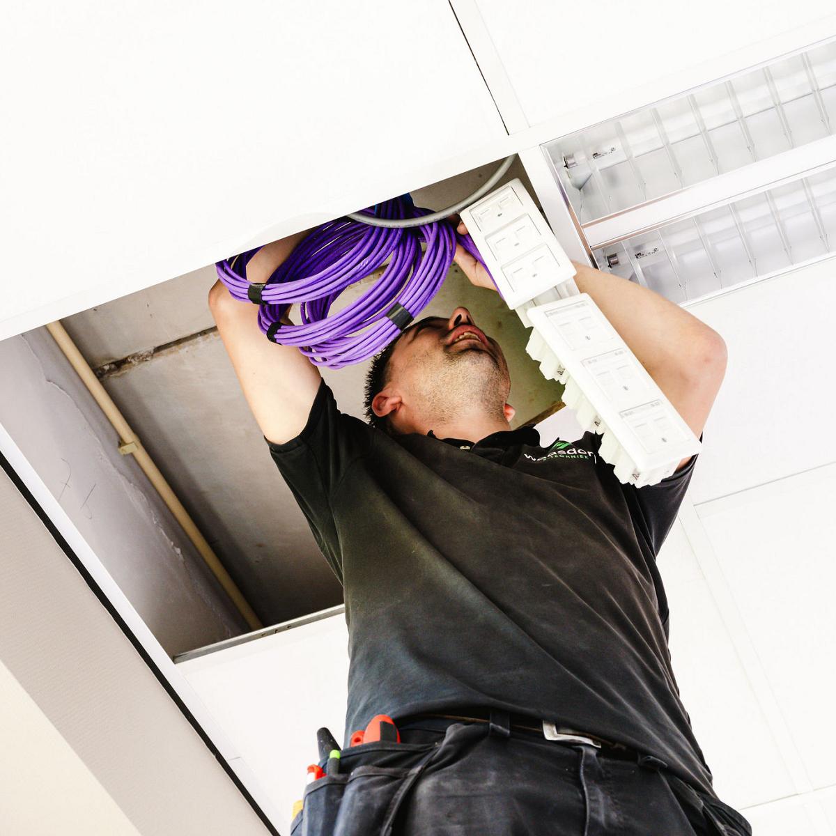Monteur werkt boven plafondplaten en trekt paarse bekabeling door voor een installatie.