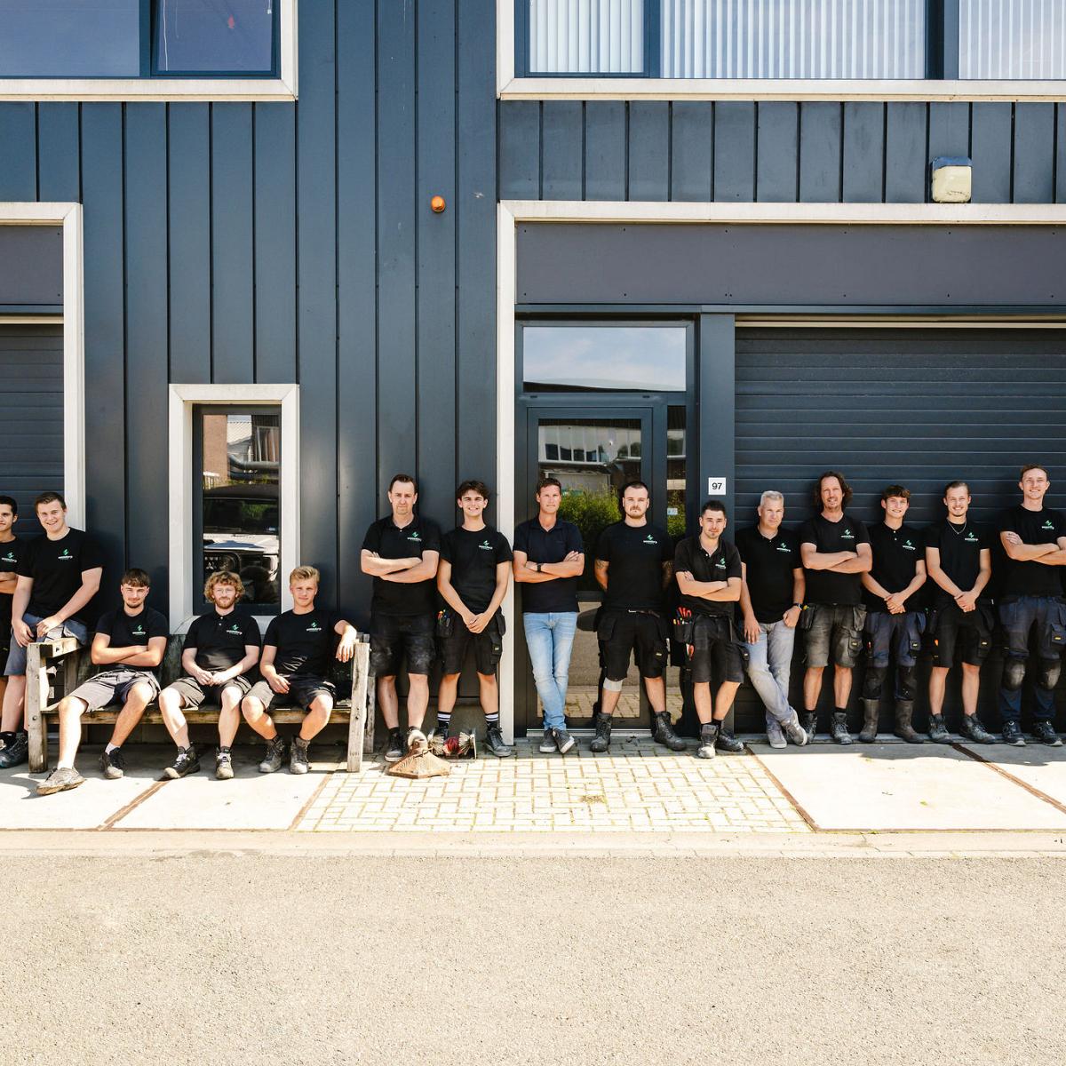 Groepsfoto van een team in zwarte werkkleding dat voor een donkerblauw gebouw staat, met meerdere garage- en raampartijen op de achtergrond.