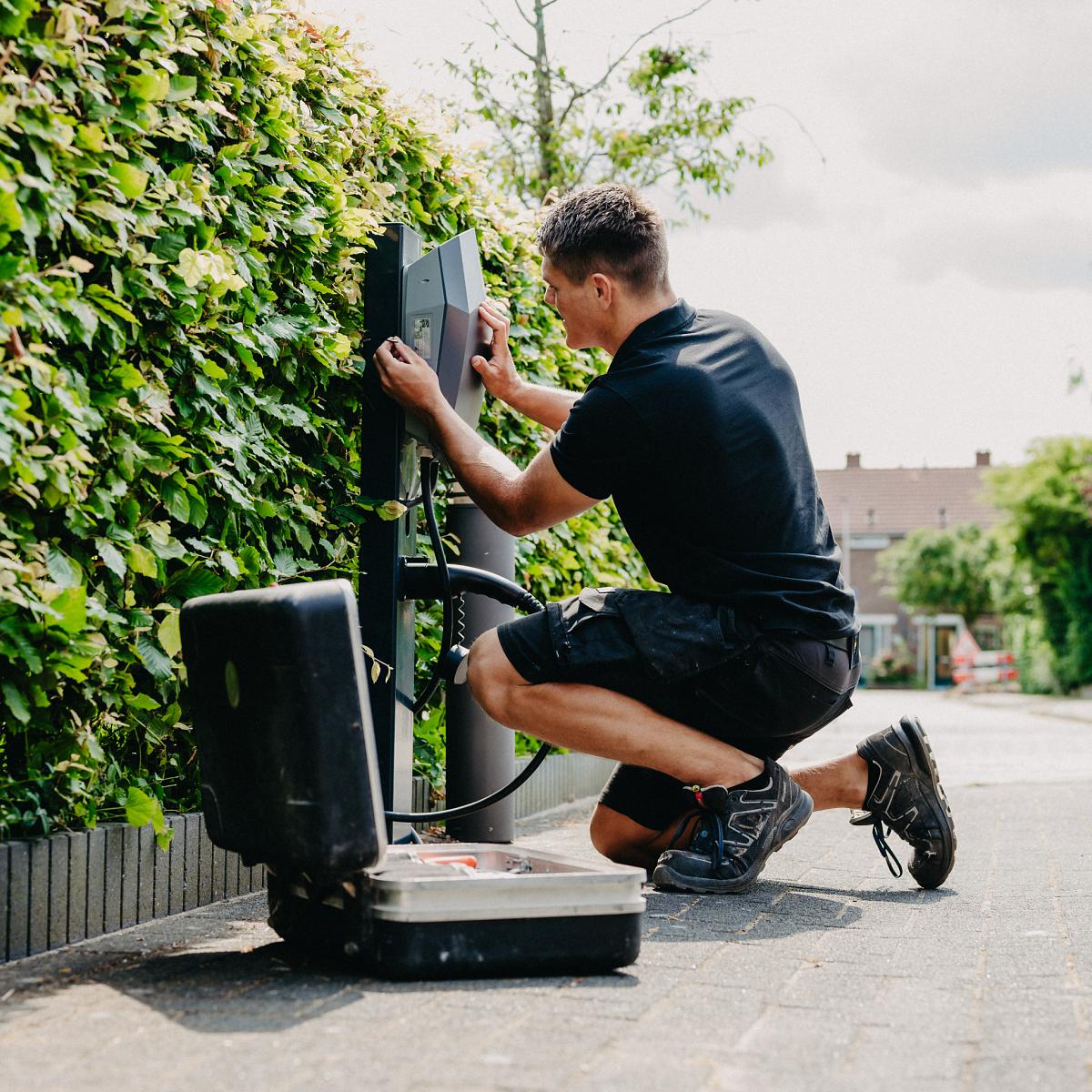 Monteur installeert een laadpaal voor elektrische voertuigen langs een groene haag, met een open gereedschapskoffer op de grond en een woonwijk op de achtergrond.