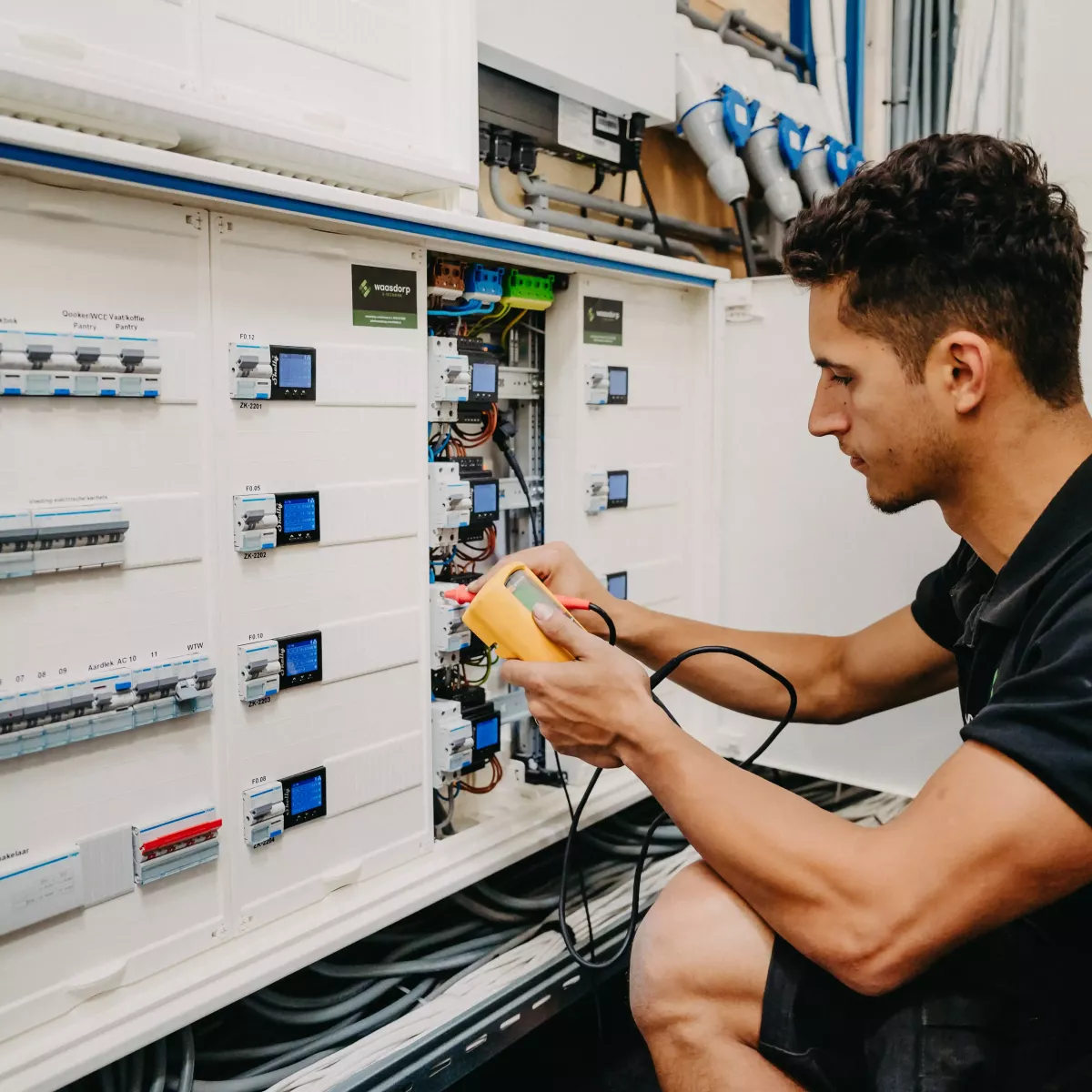 Technicus voert een meting uit in een geavanceerde elektrische verdeelkast, met meetapparatuur in de hand en zicht op bedrading en schakelaars in een technische ruimte.
