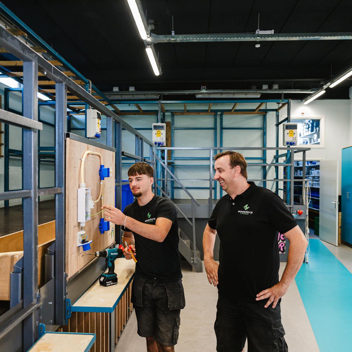 Een jonge monteur van Waasdorp E-Techniek werkt aan een elektrisch paneel, begeleid door een ervaren collega in een werkplaats met blauwe kasten en gereedschap.