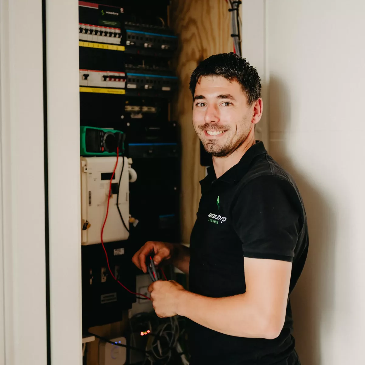 Lachende technicus werkt aan een meterkast met kabels en meetapparatuur, gekleed in Waasdorp-bedrijfskleding, in een lichte en moderne binnenruimte.