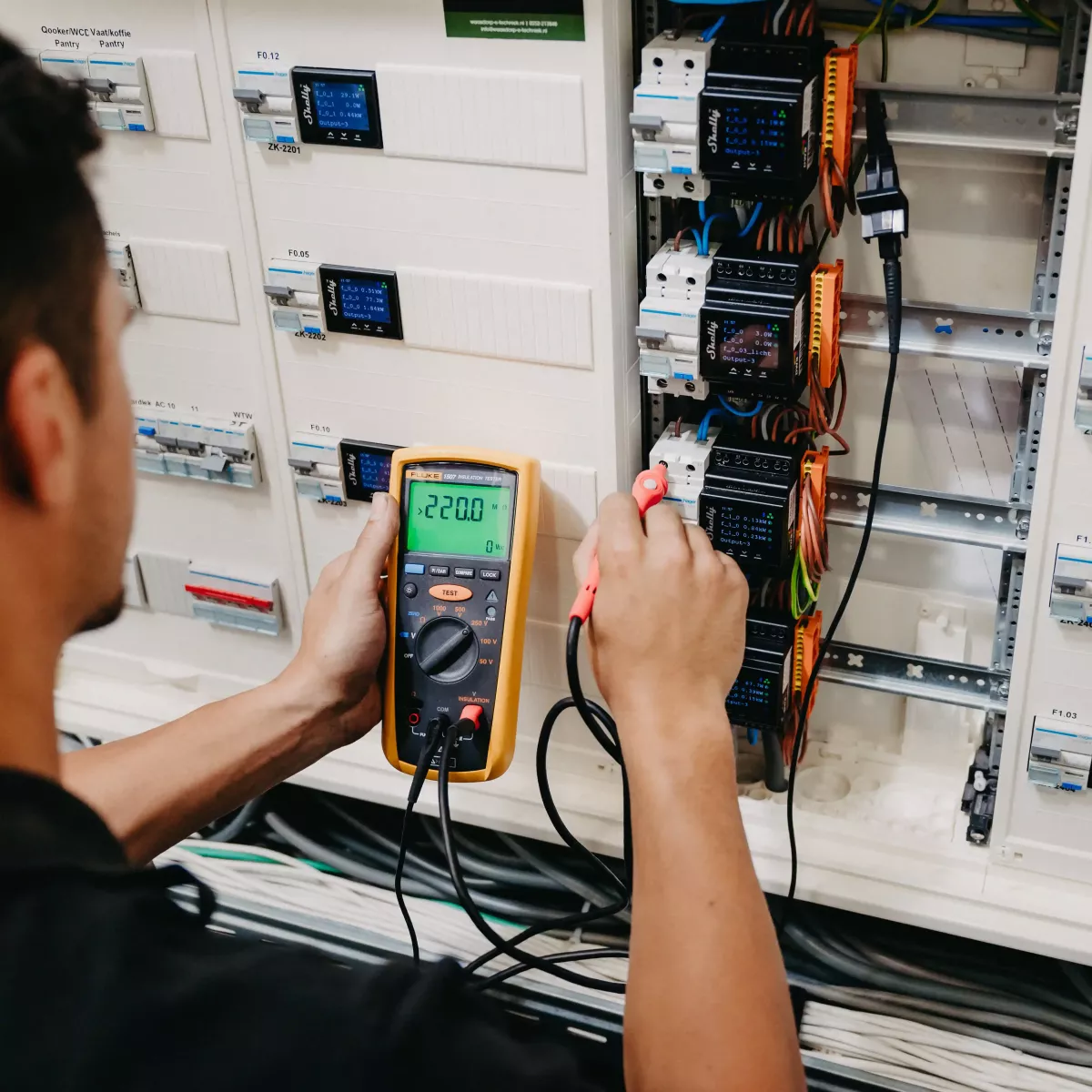 Technicus voert spanningsmeting uit met een multimeter op een elektrische installatiekast, waarbij een display 220 volt aangeeft en bedrading zichtbaar is.