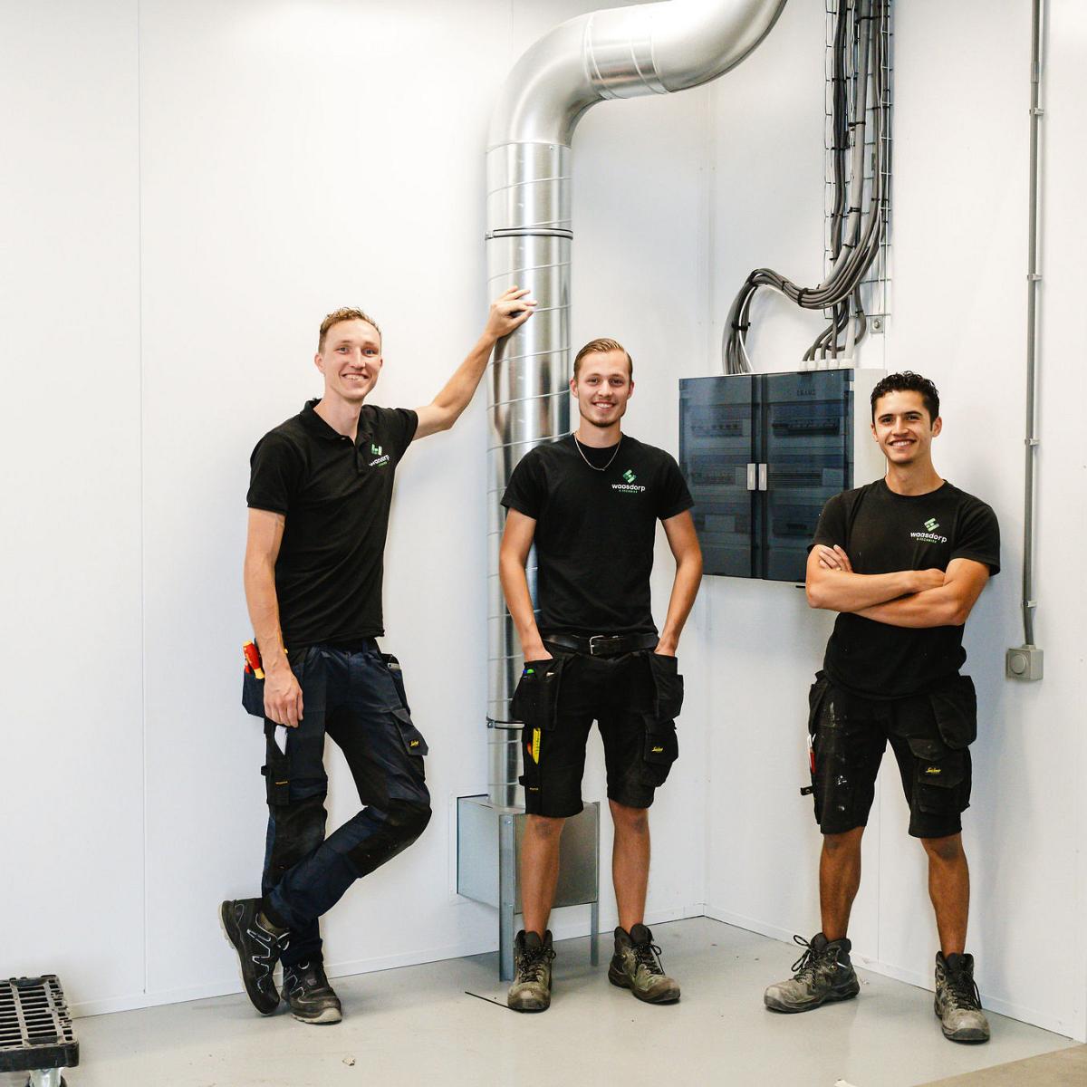 Drie monteurs van Waasdorp E-Techniek poseren lachend bij een installatie in een industriële omgeving, met zichtbare kabels en een ventilatiebuis.