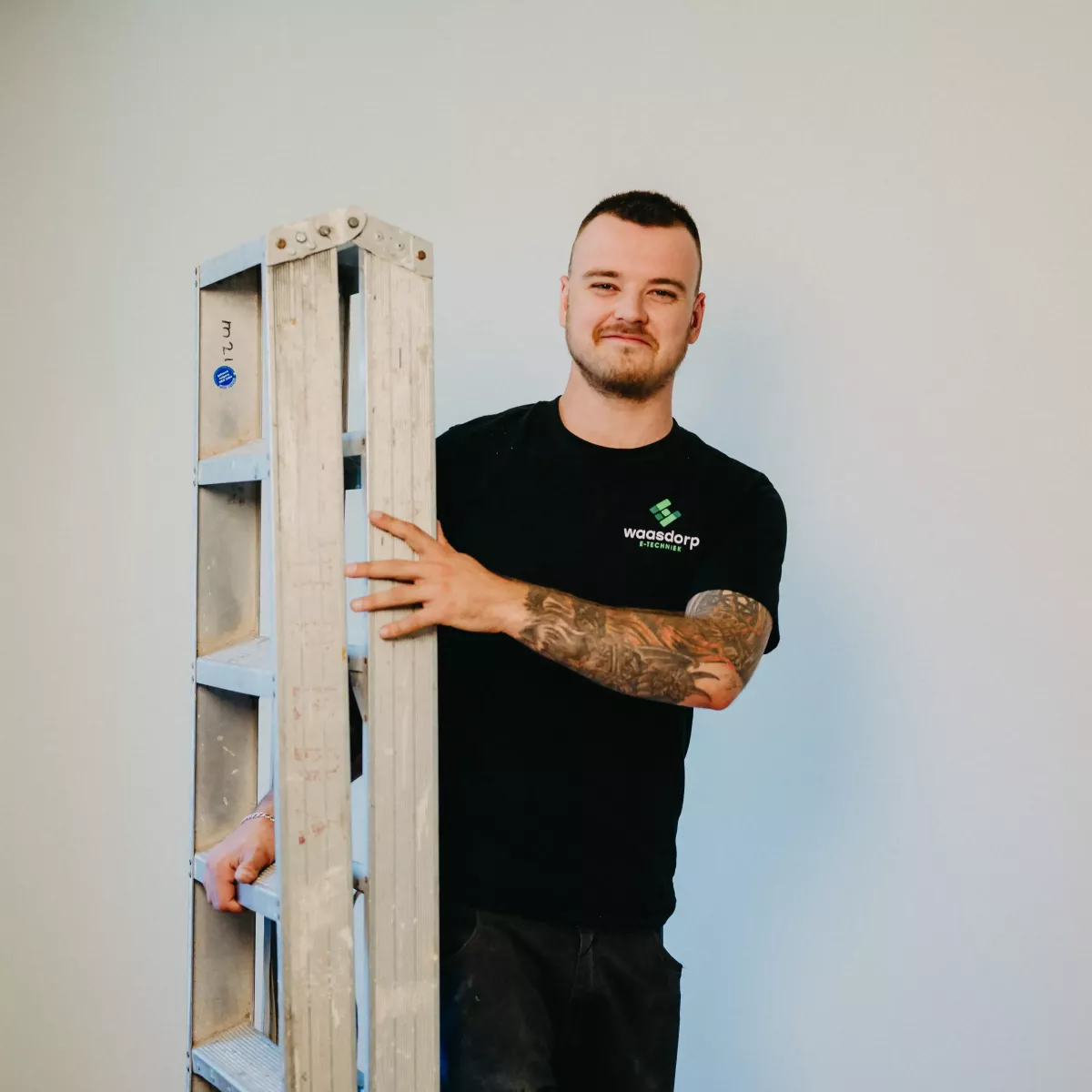 Technicus in Waasdorp-bedrijfskleding poseert met een ladder tegen een lichte achtergrond.