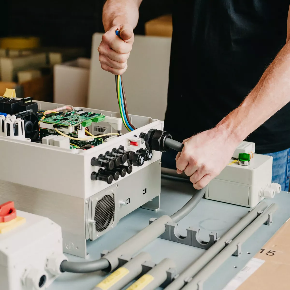 Technicus sluit een kabel aan op een open schakelkast met elektronische componenten en bedrading, geplaatst op een werkbank in een technische werkplaats.