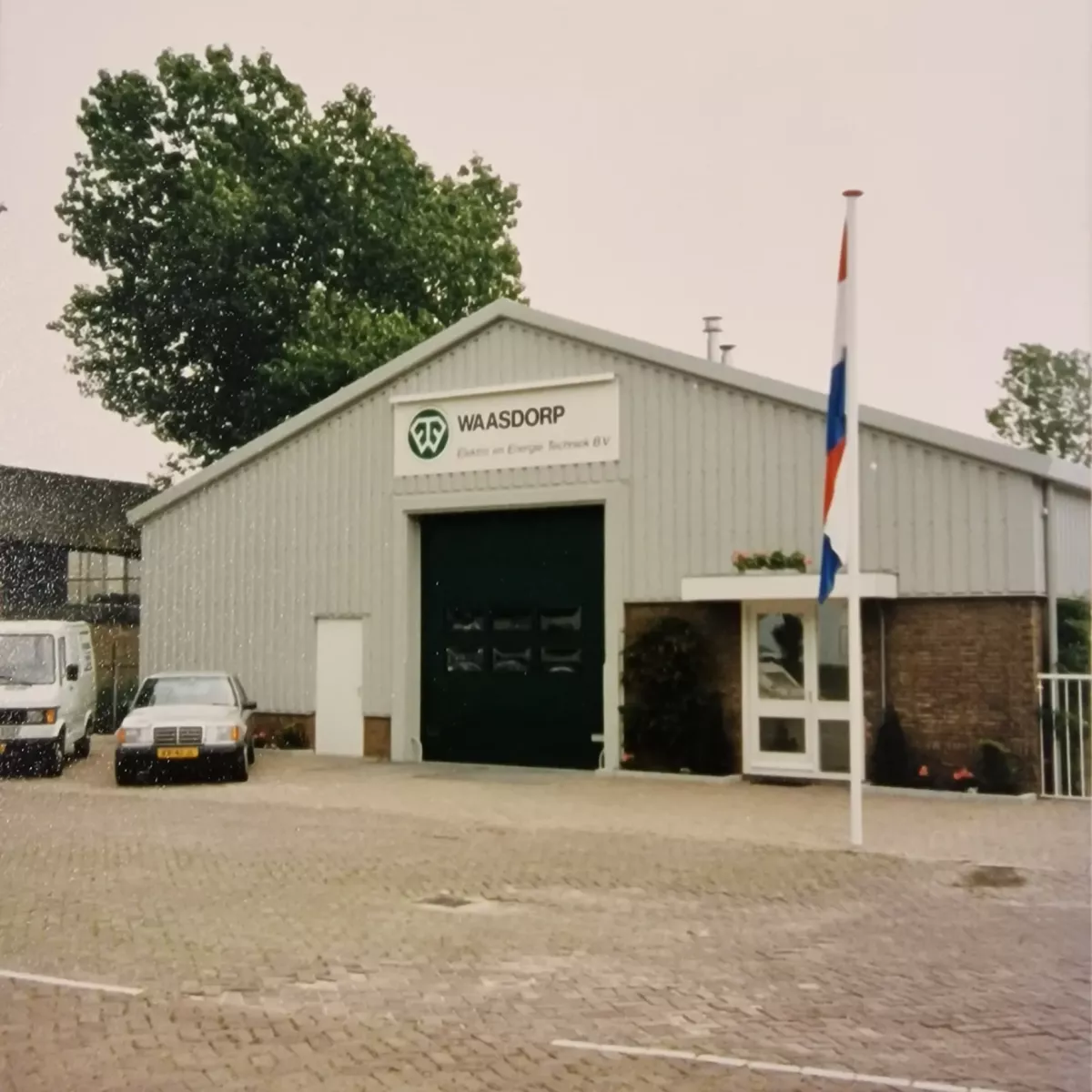 Oude bedrijfslocatie van Waasdorp Elektro en Energie Techniek B.V., met een grijs gebouw, een groene garagedeur, en een Nederlandse vlag op de voorgron