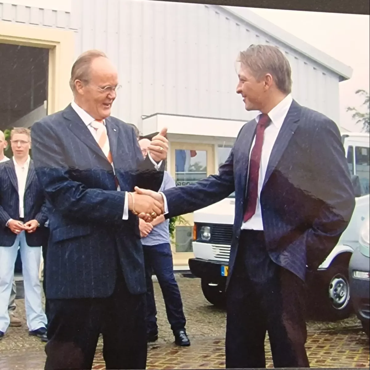 Groepsfoto bij de opening van een pand, met een handdruk tussen twee heren in pak en een groep mensen op de achtergrond.