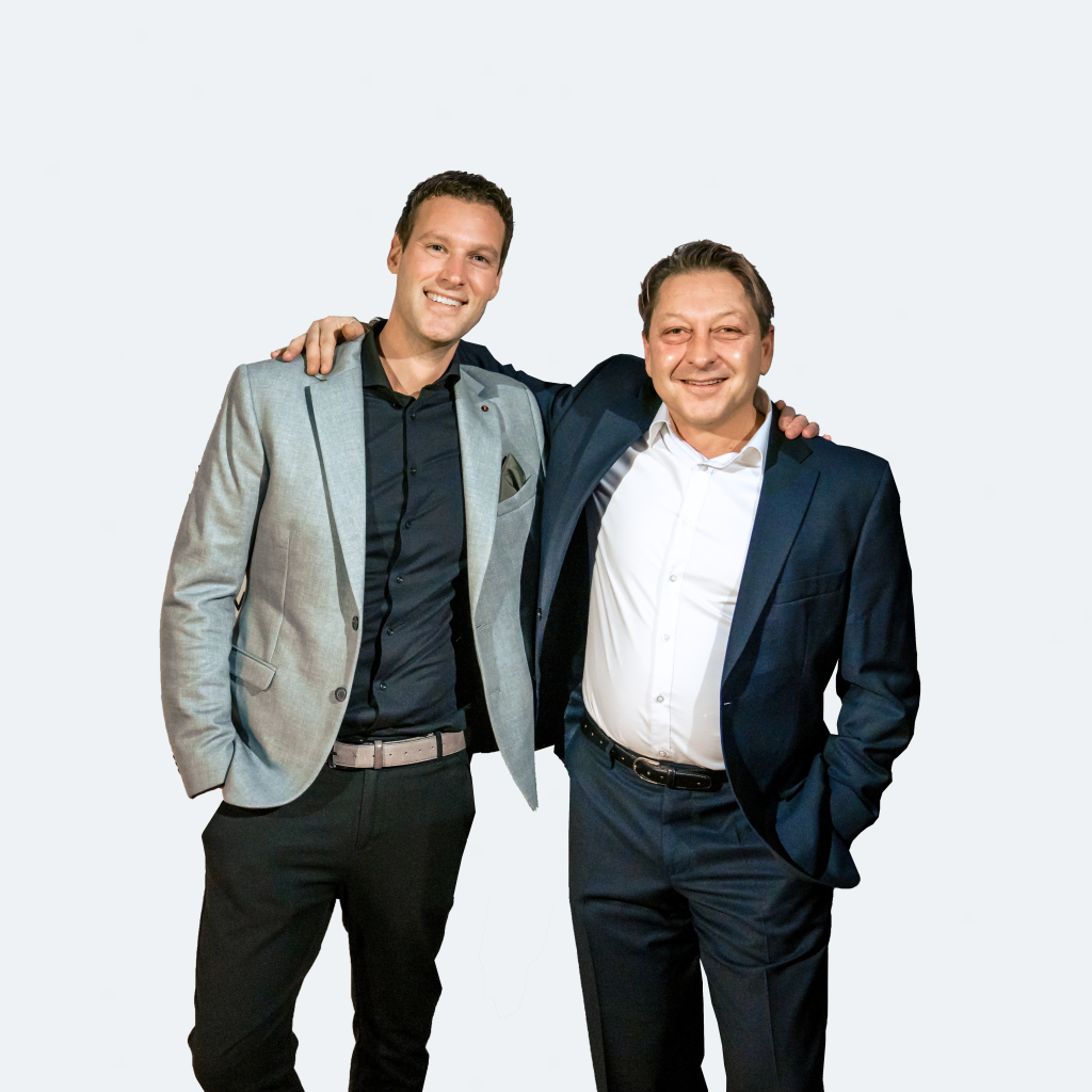 Twee mannen in nette pakken staan naast elkaar met armen om elkaars schouders, glimlachend naar de camera, tegen een witte achtergrond.