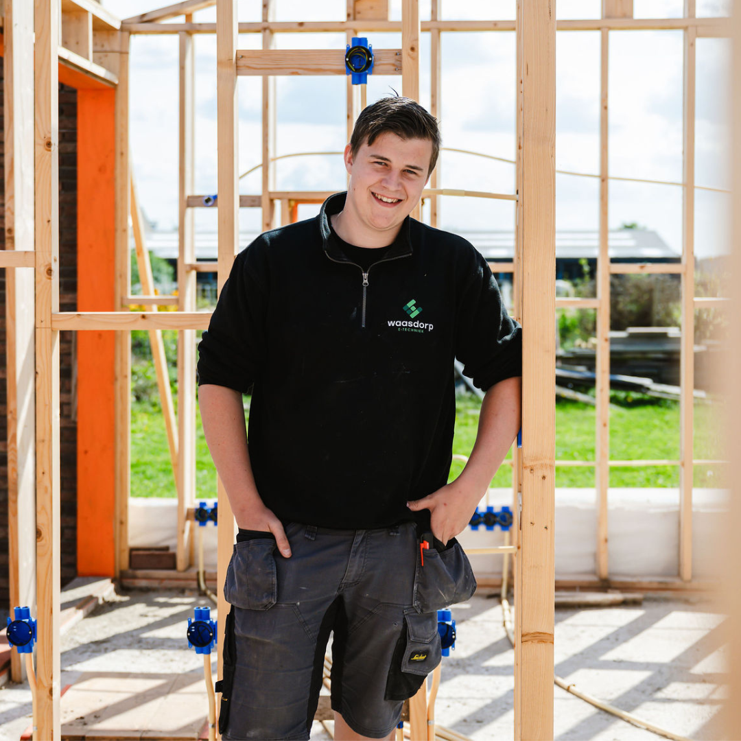 Jonge monteur in werkkleding poseert lachend tussen houten frames van een nieuwbouwwoning in aanbouw.