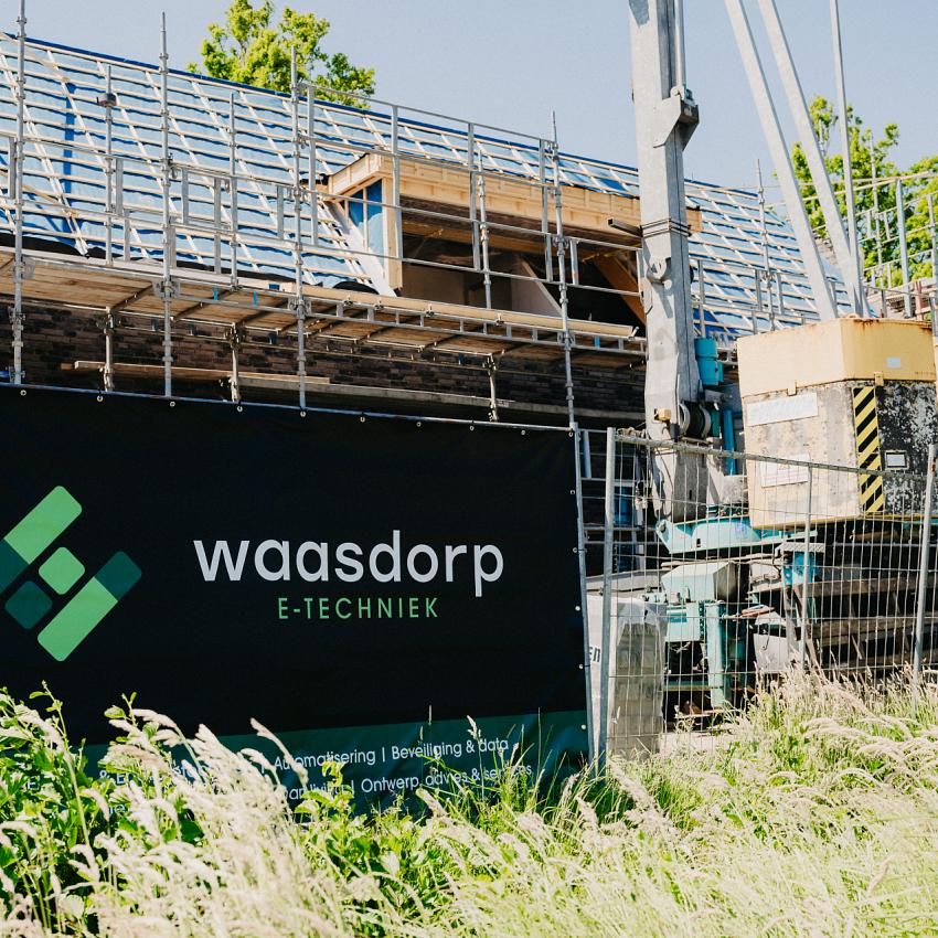 Bouwplaats met een steiger en een huis in aanbouw, omringd door groene vegetatie. Een spandoek met het logo en de naam "Waasdorp E-Techniek" hangt aan het hek.