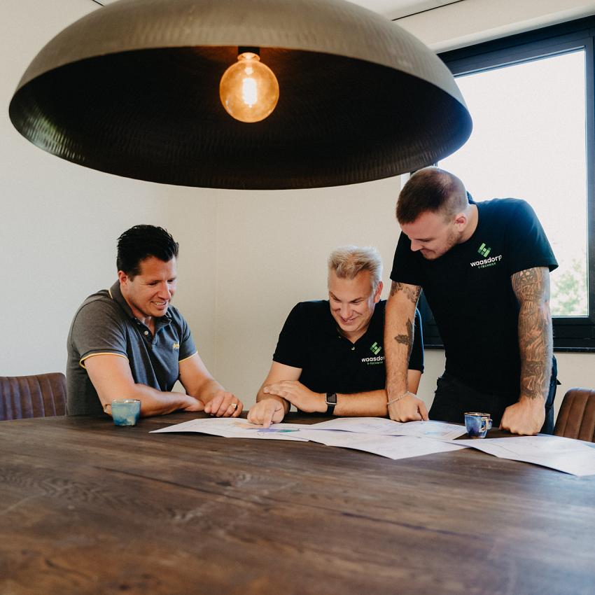 Drie mannen bespreken bouwtekeningen aan een houten tafel in een moderne vergaderruimte, verlicht door een grote hanglamp, met koffie op tafel.