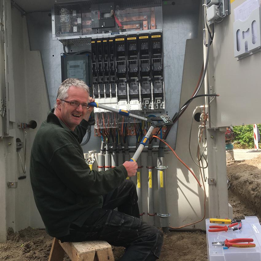 Monteur lachend bezig met het aansluiten van een groot elektrisch verdeelstation buiten, omringd door gereedschap en opengewerkte bekabeling.