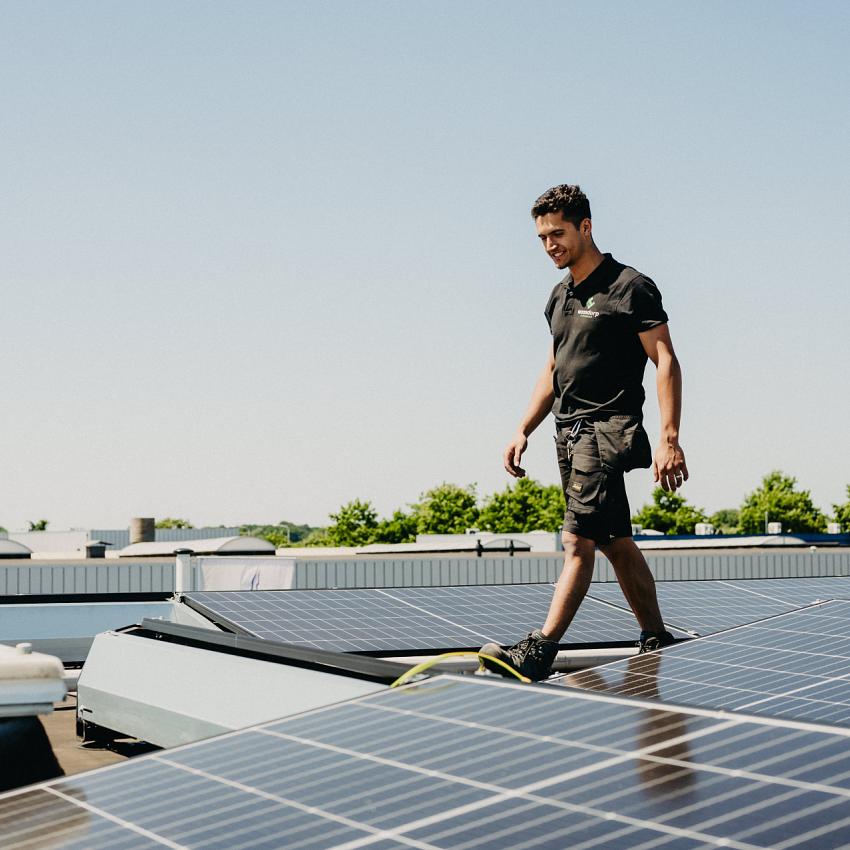 Technicus loopt over een plat dak met geïnstalleerde zonnepanelen, onder een heldere blauwe lucht, omgeven door groene bomen en industriële gebouwen.