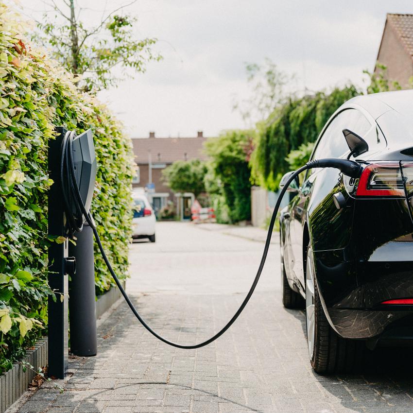 Elektrische auto aangesloten op een laadpaal naast een groene heg in een woonwijk, met rijtjeshuizen op de achtergrond en een rustige straat.