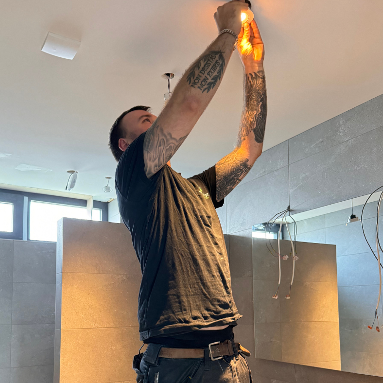 Een monteur aan het installeren van een plafondlamp in een moderne badkamer met grote grijze tegels en natuurlijk licht.