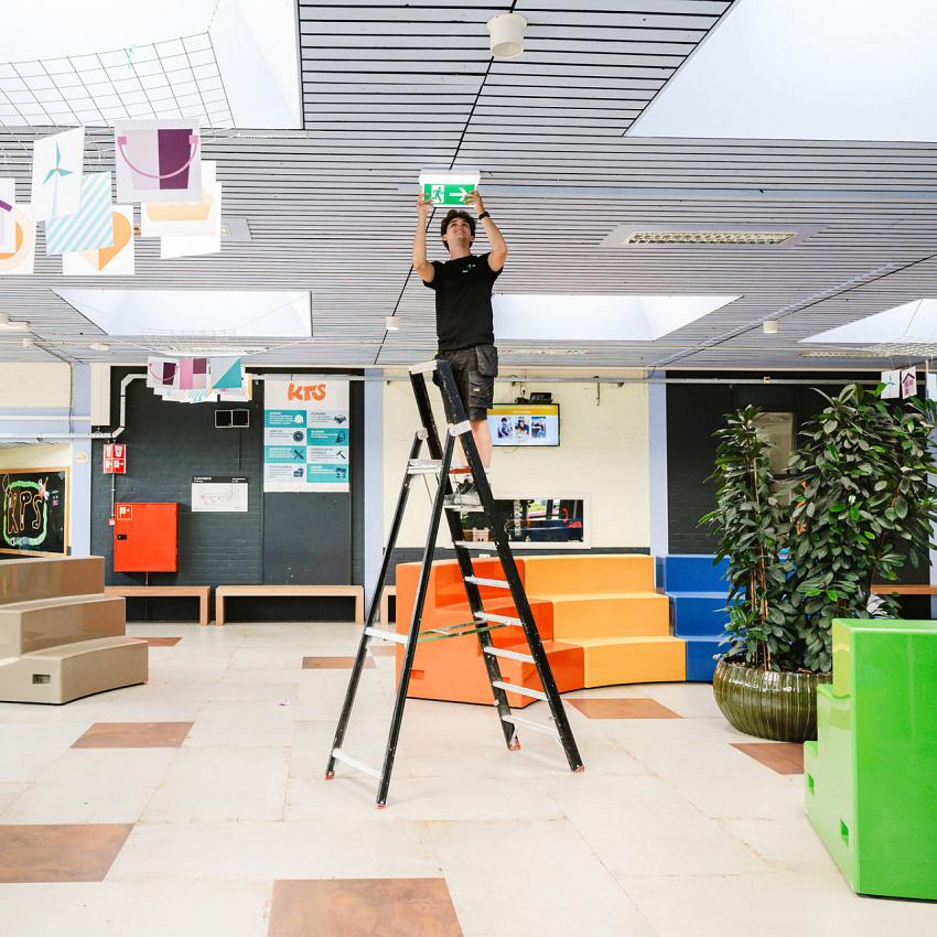 Technicus op een ladder in een open schoolruimte, bezig met het installeren of controleren van een nooduitgangbord aan het plafond. Omringd door kleurrijke decoraties en meubels.