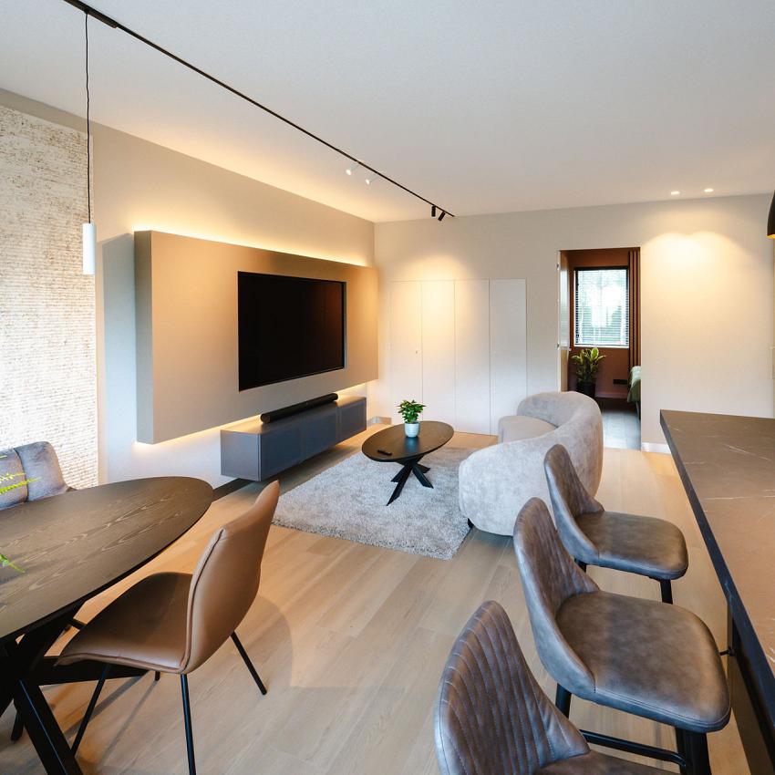 Moderne woonkamer met een strakke inrichting, voorzien van een grote tv aan de muur, een ronde eettafel met stoelen, en stijlvolle hanglampen boven een bar.