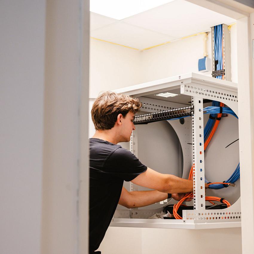 Technicus werkt in een serverkast met oranje en blauwe kabels in een technische ruimte met lichte muren en een systeemplafond.