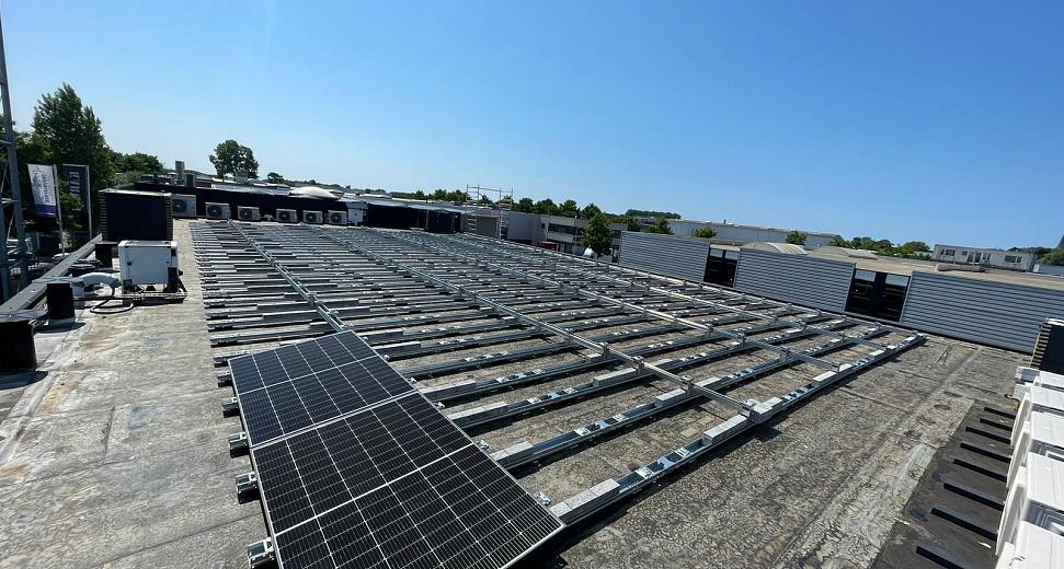 Plat dak met geïnstalleerde zonnepanelen en een metalen railsysteem, omringd door bedrijfsgebouwen en een heldere blauwe lucht op de achtergrond.