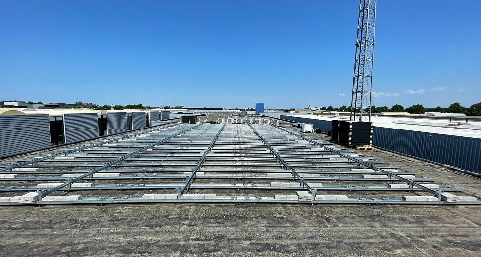 Plat dak met metalen montagerails voor zonnepanelen, omringd door industriële gebouwen en een blauwe lucht met een enkele zendmast op de achtergrond.