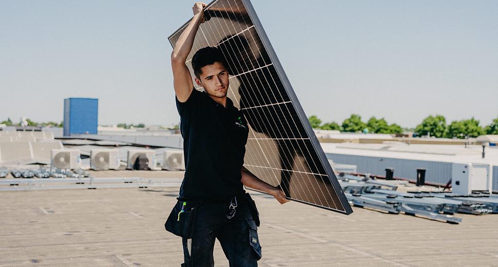Technicus draagt een zonnepaneel op een plat dak, omringd door installatiematerialen en airco-units, met een heldere blauwe lucht en groene bomen op de achtergrond.