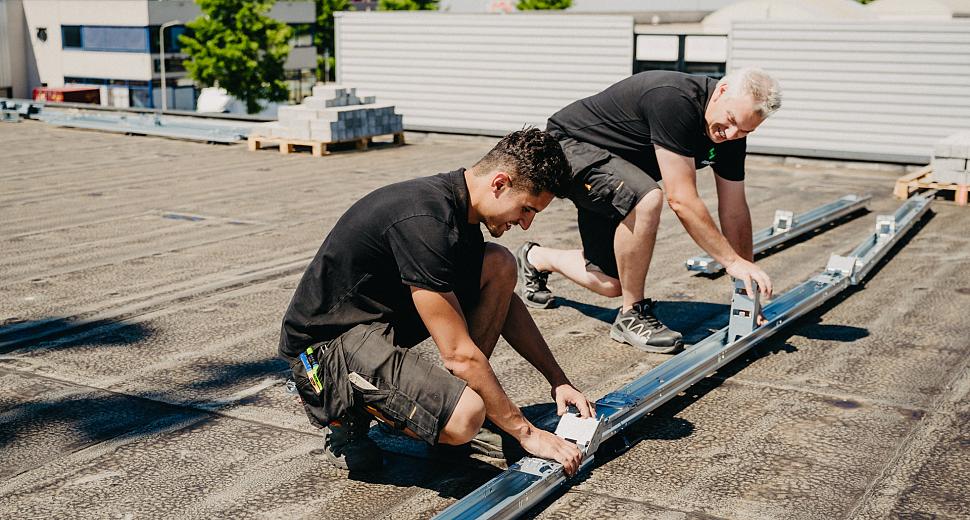 Twee monteurs bevestigen montagerails op een plat dak als voorbereiding voor de installatie van zonnepanelen, met een achtergrond van bedrijfspanden en groen.