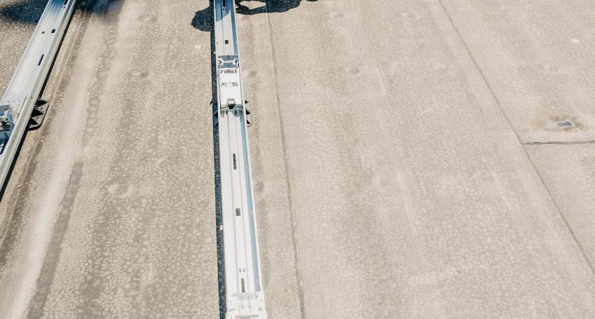 Monteur knielt op een plat dak en controleert een metalen railsysteem voor de montage van zonnepanelen, met andere installatieonderdelen op de achtergrond.