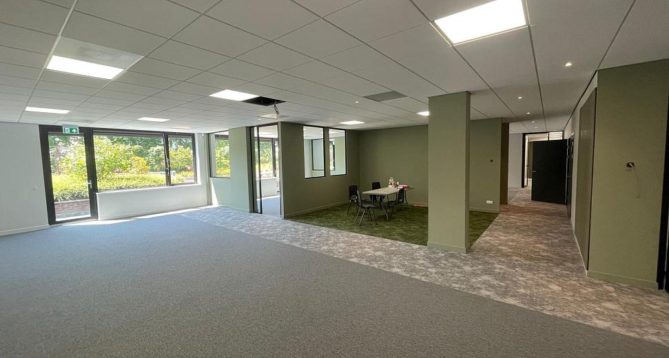 Licht en ruim kantoor met grijze vloerbedekking, groene accenten, grote ramen die uitkijken op een groene omgeving en een zithoek met een tafel en stoelen.
