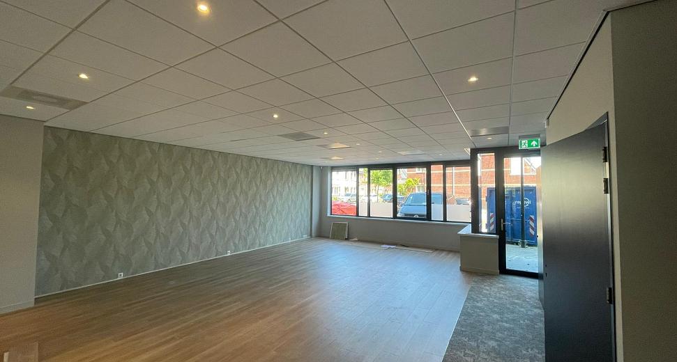 Lichte kantoorruimte met houten vloer, moderne plafondpanelen, een decoratieve wand en grote ramen die uitkijken op een stedelijke omgeving.